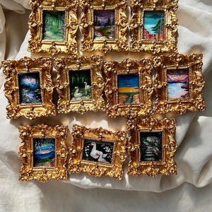 Op de afbeelding: Een verzameling miniatuur landschapsschilderijen in sierlijke gouden lijsten. De schilderijen tonen diverse scènes, waaronder bossen, zonsondergangen en kustgezichten. De lijsten hebben gedetailleerde, krullende ontwerpen en de kunstwerken staan op een witte stoffen achtergrond.