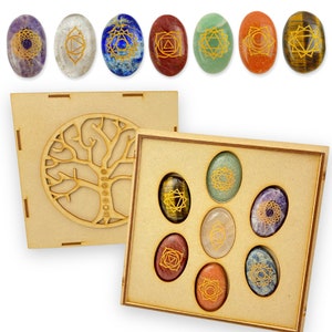 Op de afbeelding: Set van zeven chakra stenen in een houten doos. De stenen zijn ovaalvormig en hebben gouden symbolen erop. De doos heeft een levensboomontwerp op de deksel.