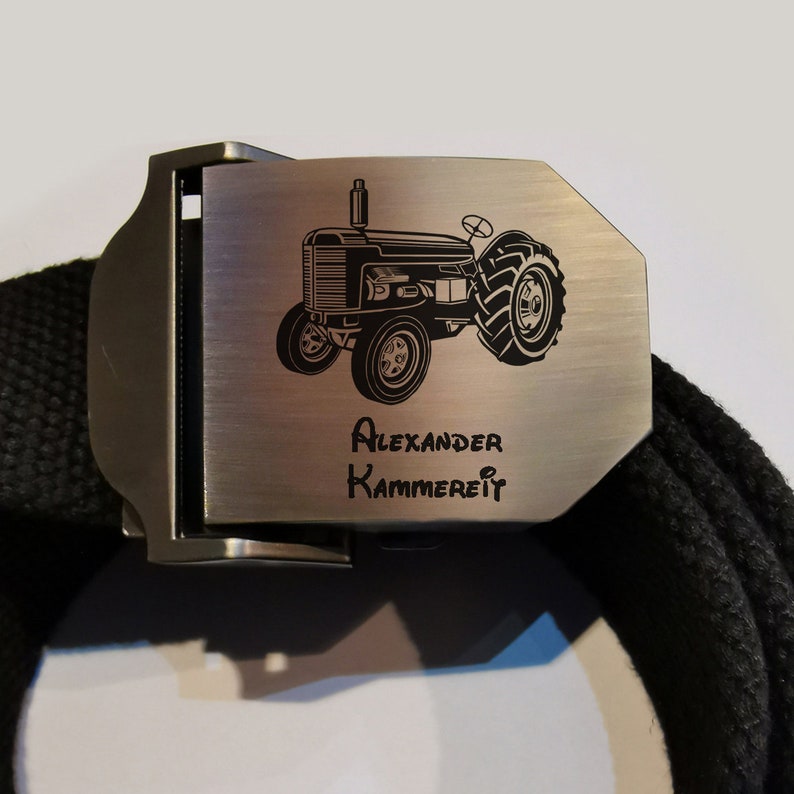 Op de afbeelding: Een zwarte canvas riem met een zilveren gesp. De gesp is gegraveerd met een afbeelding van een tractor en de tekst "Alexander Kammerer".