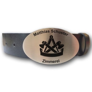 Op de afbeelding: Een zilveren gesp met een zwart-witte afbeelding van een kompas, twee bijlen en een zaag. De gesp is gegraveerd met de tekst "Matthias Schuster" en "Zimmererei".