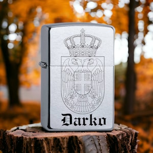 Op de afbeelding: Een zilverkleurige aansteker met een gedetailleerde gravure van een kroon en een dubbelkoppig adelaarswapen. De naam "Darko" is eronder gegraveerd. De aansteker staat op een houten oppervlak tegen een vage herfstachtergrond.