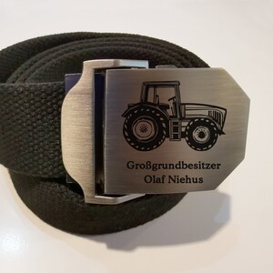 Op de afbeelding: Een zwarte canvas riem met een zilveren gesp met een tractor afbeelding en de tekst "Gro&szlig;grundbesitzer Olaf Niehus".