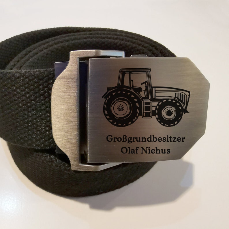 Op de afbeelding: Een zwarte canvas riem met een zilveren gesp met een tractor afbeelding en de tekst "Gro&szlig;grundbesitzer Olaf Niehus".
