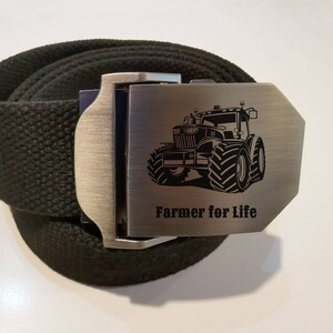 Op de afbeelding: Een zwarte canvas riem met een zilveren gesp met een tractor afbeelding en de tekst "Farmer for Life".