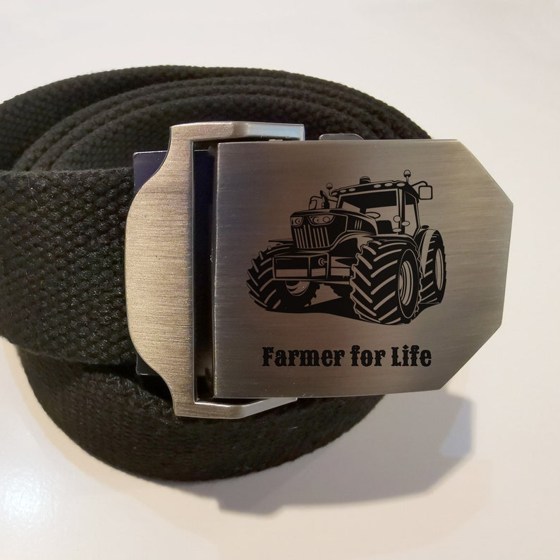 Op de afbeelding: Een zwarte canvas riem met een zilveren gesp met een tractor afbeelding en de tekst "Farmer for Life".