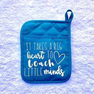 Puede incluir: Manopla de horno acolchada de color turquesa con un corazón blanco y el texto "Se necesita un gran corazón para enseñar a las mentes pequeñas".