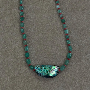 Peut inclure: Un collier composé d'un pendentif central en forme de croissant en coquille d'ormeau aux tons irisés verts et bleus, enfilé avec de petites perles vertes et de minuscules perles intercalaires rouges, terminé par un fermoir à bascule doré.