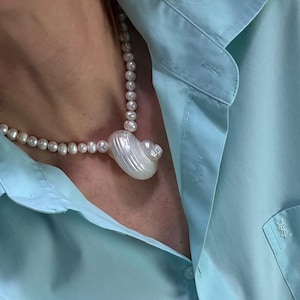 Peut inclure: Un collier de perles avec un grand pendentif en forme de coquillage blanc. Le collier est porté sur une chemise boutonnée bleu clair. La chemise a un petit logo sur la poche.