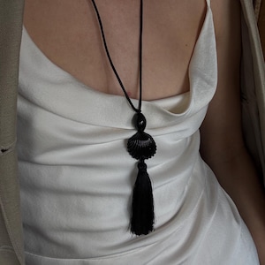 May include: A black seashell pendant necklace with a long black tassel. The necklace is worn over a white satin dress and a tan blazer.