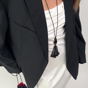 Puede incluir: Un blazer negro sobre un vestido lencero de satén blanco, combinado con un collar de cordón largo con una borla de cuentas negras. Se sostiene una pequeña bolsa negra con cordón y forro rojo, y se llevan sandalias negras de punta abierta.