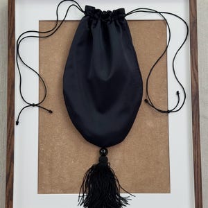 May include: A black drawstring pouch made of a smooth, satin-like fabric. The bag features a gathered top closure with thin black cord strings and a decorative black tassel hanging from the bottom, accented by a single round black bead.
