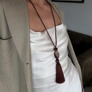 Puede incluir: Un collar largo con un cordón trenzado de color burdeos oscuro, una cuenta redonda lisa, un colgante de piedra veteada de forma ovalada y una borla a juego en color burdeos. El collar se lleva sobre un top de satén blanco y una americana beige.