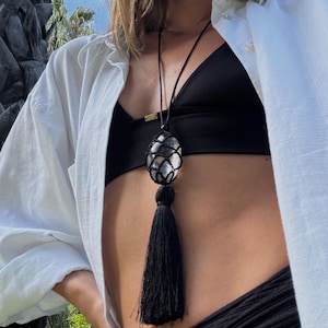 May include: A black necklace with a large, oval-shaped pendant encased in a woven net. The pendant has a silver and clear stone. A long black tassel hangs below the pendant. The necklace is worn against a black top and white shirt.