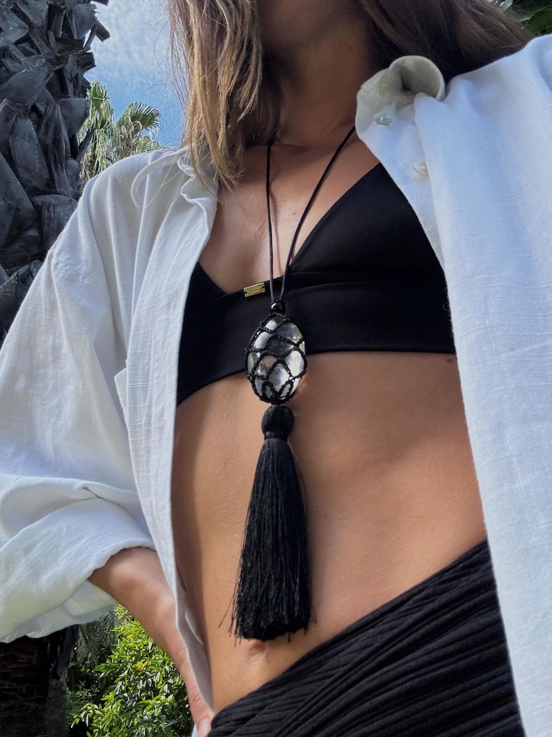 May include: A black necklace with a large, oval-shaped pendant encased in a woven net. The pendant has a silver and clear stone. A long black tassel hangs below the pendant. The necklace is worn against a black top and white shirt.