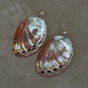 Peut inclure: Une paire de boucles d'oreilles pendantes composées de grandes coquilles d'ormeau ovales aux motifs irisés et tourbillonnants de brun, de crème et de blanc nacré. Chaque coquille présente une rangée de petits trous et un clou rond transparent.