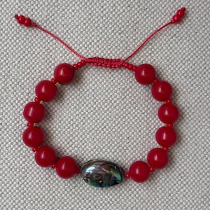 Puede incluir: Una pulsera de cuentas rojas con una cuenta central ovalada de concha de abulón iridiscente. La pulsera está hecha con cuentas rojas redondas y lisas, e incluye un cierre deslizante ajustable con dos cordones rojos terminados en cuentas.