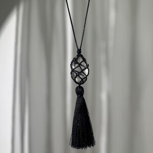May include: A black necklace featuring a clear, oval-shaped stone encased in a woven net. A black tassel hangs below the stone. The necklace is suspended by a thin, black cord against a soft, neutral background.