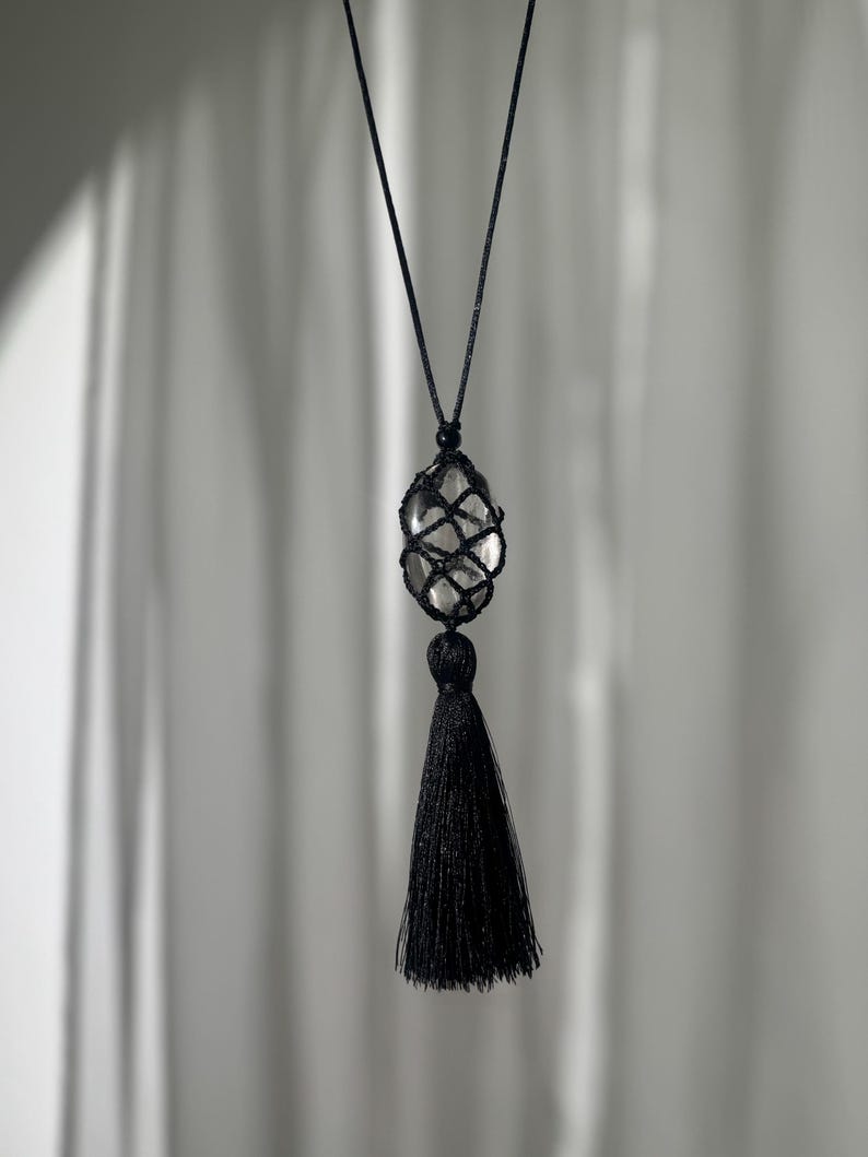 May include: A black necklace featuring a clear, oval-shaped stone encased in a woven net. A black tassel hangs below the stone. The necklace is suspended by a thin, black cord against a soft, neutral background.