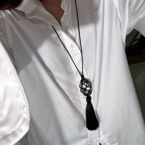 May include: A black necklace with a large, clear crystal encased in a woven net design. The necklace features a long black tassel pendant. The necklace is worn over a white button-down shirt.