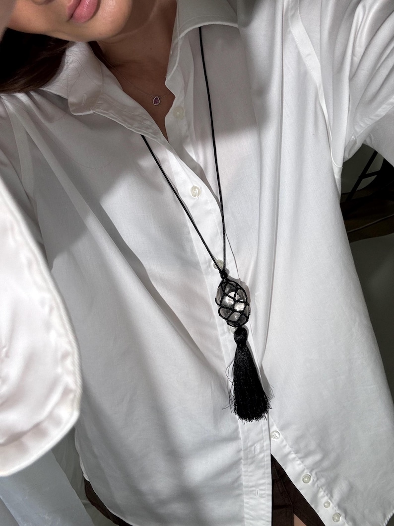 May include: A black necklace with a large, clear crystal encased in a woven net design. The necklace features a long black tassel pendant. The necklace is worn over a white button-down shirt.
