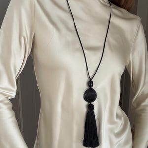 Op de afbeelding: Een zwarte ketting met een schelpenhanger en kwast, gedragen over een crèmekleurige coltrui met lange mouwen. De hanger heeft een ronde, zwarte kraal boven een schelpmotief.