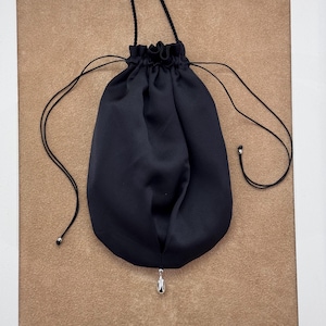 Peut inclure: Une pochette à cordon noire en tissu lisse avec une fermeture froncée sur le dessus. Le sac présente de fines cordes noires et une breloque argentée en forme de goutte d'eau suspendue au centre, avec de petites perles argentées aux extrémités.