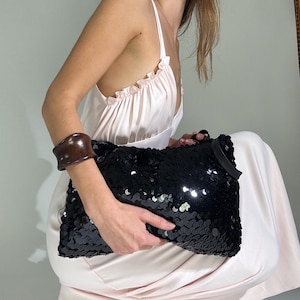 May include: A pale pink sleeveless dress with a ruffled neckline, a dark brown wide cuff bracelet, and a rectangular black sequined clutch bag. The ensemble is paired with black strappy heeled sandals, creating a contrast between the light fabric.