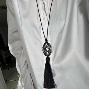 May include: A long black necklace with a large, oval-shaped, clear stone encased in a black net. The necklace has a long black tassel at the bottom. The necklace is worn over a white button-down shirt.