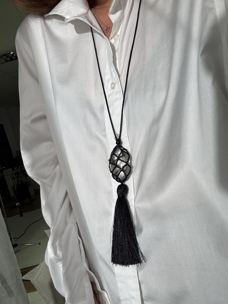 May include: A long black necklace with a large, oval-shaped, clear stone encased in a black net. The necklace has a long black tassel at the bottom. The necklace is worn over a white button-down shirt.