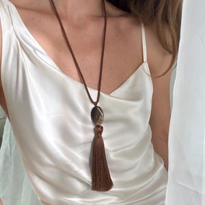 Könnte beinhalten: Eine lange Halskette mit einer braunen gedrehten Kordel, einem glatten ovalen braun-grauen Steinanhänger und einer langen braunen Stoffquaste. Die Kette wird über einem weißen Satin-Top mit Wasserfall-Ausschnitt und dünnen Trägern getragen.
