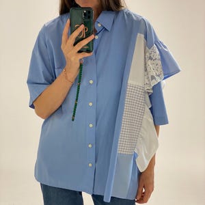 Puede incluir: Una camisa azul claro de botones con un panel a cuadros blanco y azul, un panel blanco con ribete de encaje y un panel azul claro. La camisa tiene cuello y mangas cortas.