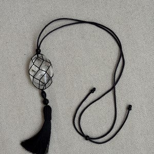 Puede incluir: Un collar con colgante de cristal de cuarzo transparente. El cristal está encerrado en una red negra tejida, con una borla negra y un cordón negro ajustable. El colgante tiene forma ovalada y cuelga de un cordón largo y ajustable.