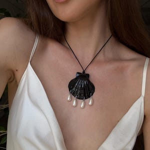 Puede incluir: Un collar con colgante de concha negra con perlas de imitación colgantes. El colgante es de color negro brillante y cuelga de un fino cordón negro. El collar se lleva sobre una blusa de satén blanca.