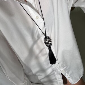 May include: A black macrame necklace with a spherical, crystal-like pendant and a black tassel. A smaller silver necklace with a pink gemstone is also visible. The necklaces are worn over a white button-down shirt.