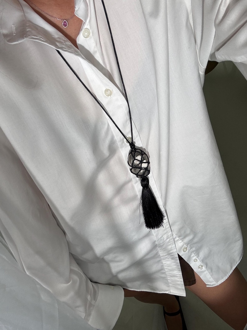 May include: A black macrame necklace with a spherical, crystal-like pendant and a black tassel. A smaller silver necklace with a pink gemstone is also visible. The necklaces are worn over a white button-down shirt.