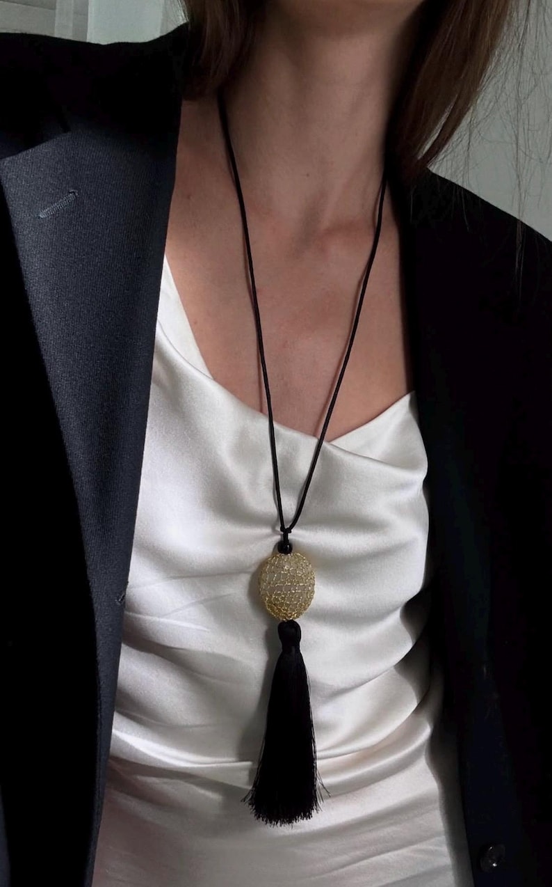 May include: A long necklace with a gold-colored, textured oval pendant and a black tassel. The pendant hangs from a black cord. The necklace is worn over a white satin top and a black blazer.