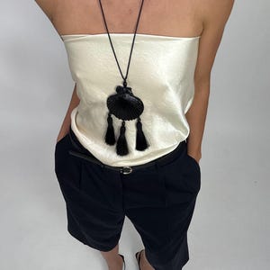 Pode incluir: Um colar com pingente de concha preta com três borlas é usado sobre um top de tubo creme. A modelo usa shorts pretos, um cinto preto e sapatilhas pretas de bico fino. A roupa é simples e elegante.