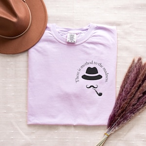 May include: A light purple t-shirt featuring a black hat, mustache, and pipe design with the text "There is method to the madness." A brown hat is visible in the upper left corner. The shirt is a Comfort Colors brand.