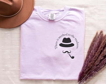 May include: A light purple t-shirt featuring a black hat, mustache, and pipe design with the text "There is method to the madness." A brown hat is visible in the upper left corner. The shirt is a Comfort Colors brand.