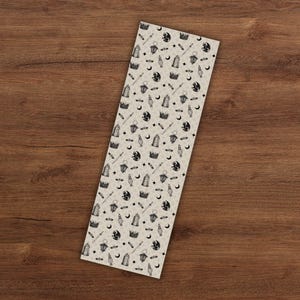 May include: A rectangular bookmark with a repeating pattern of black and white fantasy-themed illustrations. The design includes crowns, moons, swords, lanterns, and stars on a beige background. The bookmark is displayed on a wooden surface.