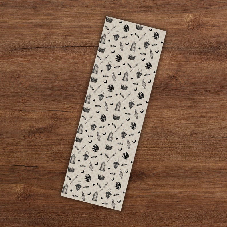 May include: A rectangular bookmark with a repeating pattern of black and white fantasy-themed illustrations. The design includes crowns, moons, swords, lanterns, and stars on a beige background. The bookmark is displayed on a wooden surface.