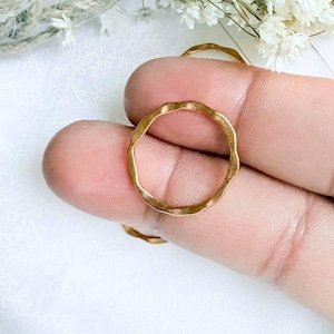 May include: A thin gold ring with a wavy, undulating band design. The ring features a smooth, polished metallic finish and a circular shape. The band has a consistent thickness throughout its circumference, creating a simple and clean aesthetic.