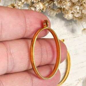 May include: A gold-toned oval pendant bezel with a smooth, metallic finish and a small circular loop at the top for attachment. The frame is hollow in the center, designed to hold a cabochon or insert, and features a simple, clean, and open design.