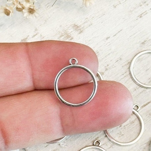 May include: A silver-toned metal circular charm featuring a thin, smooth frame and a small jump ring at the top for attachment. The charm has a hollow center and a polished finish, presented as a single piece held up for display among others.