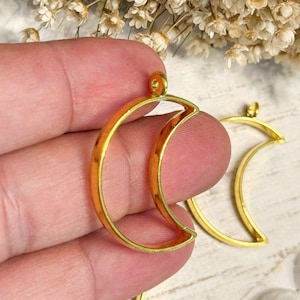 May include: Two gold-colored metal crescent moon pendants with open centers and small loops at the top for attachment. The pendants have a smooth, reflective finish and a curved shape, designed as frames for jewelry making or decorative craft projects.