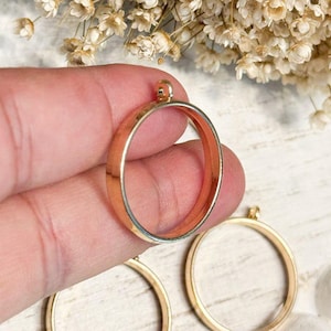 May include: A gold-toned circular pendant bezel with a small loop at the top for attachment. The metal frame is smooth and polished, designed to hold a circular insert. The object is held in a position that displays its round shape and depth.