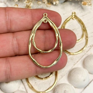 May include: A gold-toned metal jewelry component featuring a double teardrop frame design with a textured, organic surface. The piece includes a small loop at the top for attachment, showcasing a layered, open-work structure for crafting.