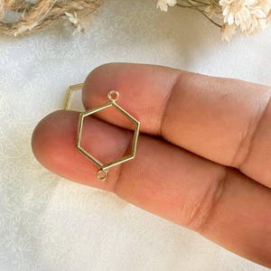May include: Gold-toned hexagonal pendant with two jump rings. The pendant is open and can be used to hold a charm or other embellishment.