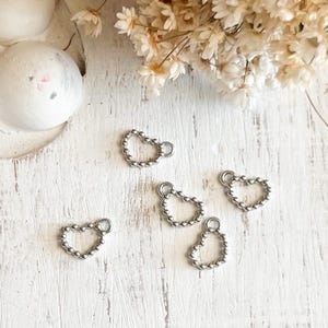 May include: Five silver-colored heart-shaped charms with a textured, beaded design. Each charm has a small loop at the top. The charms are scattered on a white, textured surface, with dried flowers in the background.