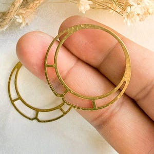 May include: Two gold-colored, open-circle shaped earrings with a geometric design. The earrings are held in a person's hand, with a soft, neutral background. The earrings have a hammered texture and are likely for jewelry making.